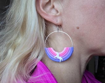 May include: Silver hoop earrings with a rainbow design in shades of pink, purple, and blue. A white unicorn silhouette is centered on the rainbow. The earrings are a whimsical accessory.