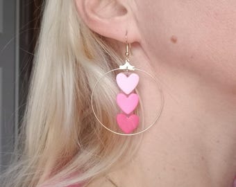 May include: Gold hoop earrings with three pink heart charms, each heart is a different shade of pink, from light to dark.