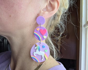 May include: A pair of whimsical, dangling earrings. Each earring features three lavender-colored circles connected by silver links. The circles are decorated with hearts, rainbows, and a white unicorn. The earrings are a fun, colorful accessory.