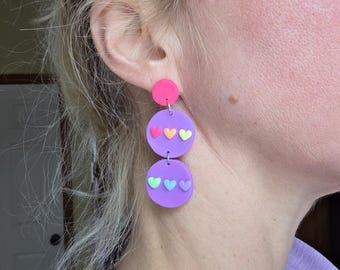 May include: A pair of colorful earrings with a pink top circle and two larger purple circles below. Each purple circle has three small heart shapes in different colors. The earrings are a fun and playful accessory.