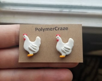 May include: A pair of white chicken earrings with red combs and yellow feet. The earrings are displayed on a brown card with the text "PolymerCraze".