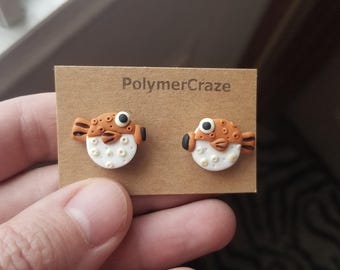 May include: A pair of handmade polymer clay earrings featuring a pufferfish design. The earrings are orange and white with black eyes. The earrings are on a card with the text "PolymerCraze".