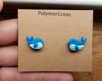 May include: A pair of blue whale stud earrings. Each earring features a blue whale with a white belly and a small, curved tail. The earrings are displayed on a brown card with the text "PolymerCraze".