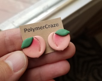 May include: A pair of peach-colored polymer clay earrings with green leaves. The earrings are shaped like peaches and have a realistic look. The earrings are on a card that says "PolymerCraze".