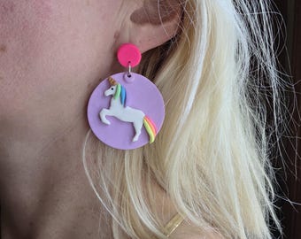 May include: A pair of whimsical unicorn earrings. Each earring features a white unicorn with a rainbow mane and tail, set against a lavender circular backdrop. The earrings are connected to a small, bright pink stud.