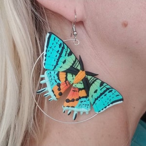 May include: A pair of silver hoop earrings with a colorful butterfly charm. The butterfly is made of polymer clay and has a blue, green, orange, and black design.