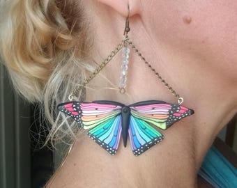 May include: A pair of butterfly earrings. The earrings feature a colorful butterfly design with rainbow wings and black detailing. The butterfly is suspended from a chain and a beaded strand. The earrings are a unique accessory.