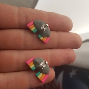 May include: Rainbow-shaped stud earrings. Each earring features a gray cloud with a small, clear gem and a colorful rainbow design in pink, orange, yellow, green, and blue. The earrings are small and delicate.