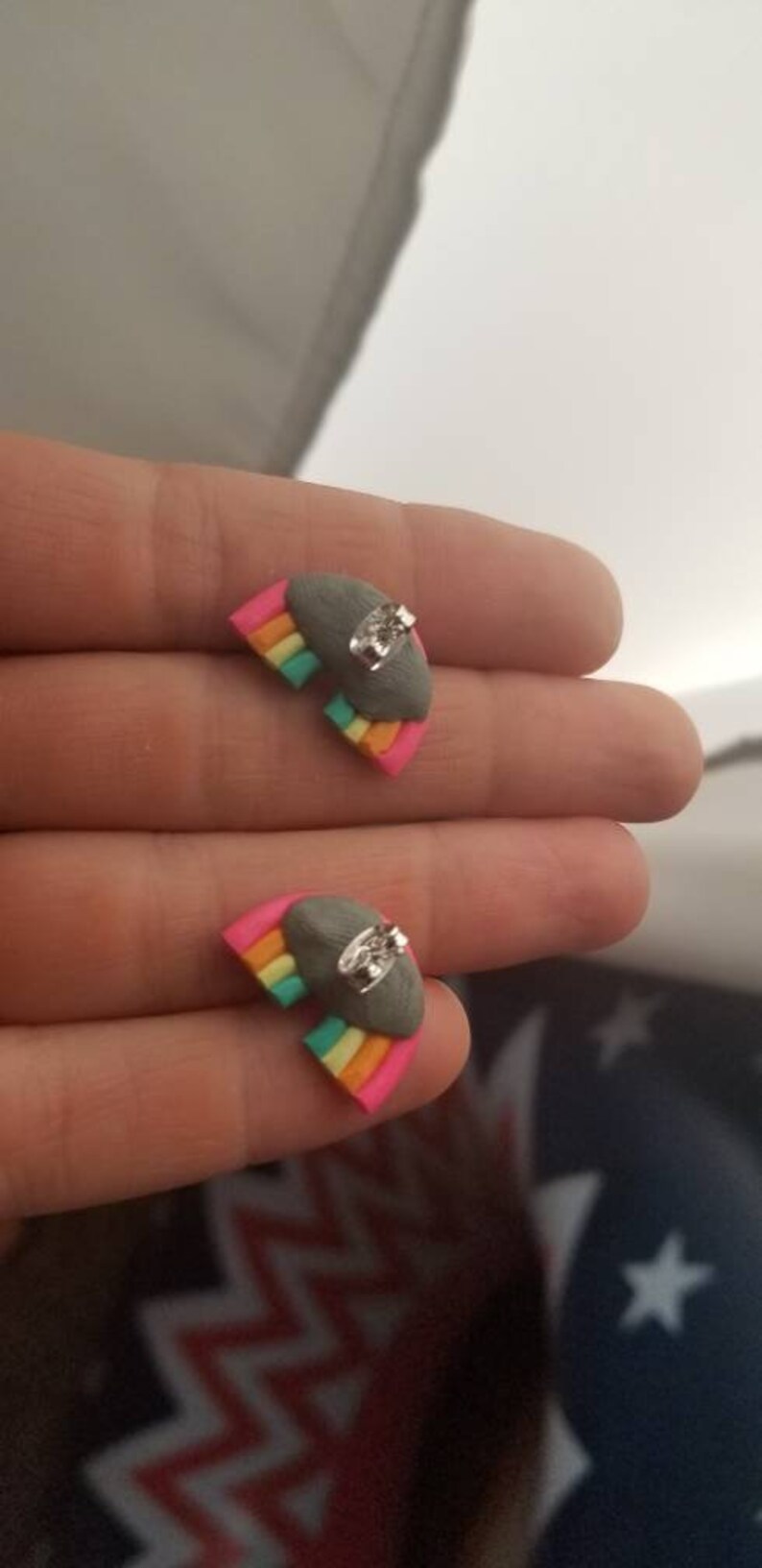 May include: Rainbow-shaped stud earrings. Each earring features a gray cloud with a small, clear gem and a colorful rainbow design in pink, orange, yellow, green, and blue. The earrings are small and delicate.