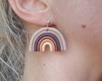 May include: A pair of rainbow-colored clay earrings. The earrings are shaped like a rainbow with five different colors: light brown, orange, yellow, dark brown, and pink. The earrings are attached to silver hooks.
