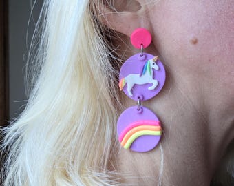 May include: A pair of whimsical, dangling earrings. The earrings feature a pink stud, a purple disc with a white unicorn, and a lower purple disc with a rainbow design. The unicorn has a colorful mane and tail. The rainbow has yellow, orange, and pink stripes.