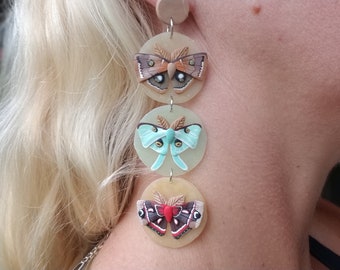 May include: A set of three dangle earrings featuring a layered design with three different butterfly motifs. The top butterfly is brown with orange accents, the middle butterfly is light blue with yellow accents, and the bottom butterfly is red with black and white accents. The butterflies are all set against a cream-colored background.