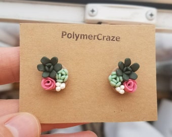 May include: A pair of polymer clay earrings featuring a succulent plant design with green leaves, pink flowers, and white accents. The earrings are displayed on a brown card with the text "PolymerCraze".