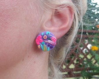 May include: Purple and pink polymer clay earrings with a colorful abstract design. The earrings are round and feature a variety of shapes and colors, including green, blue, and yellow.