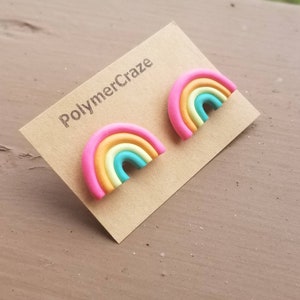 May include: A pair of rainbow-shaped stud earrings. Each earring features a pink outer band, followed by bands of orange, yellow, and turquoise. The earrings are displayed on a brown card with the text "PolymerCraze".