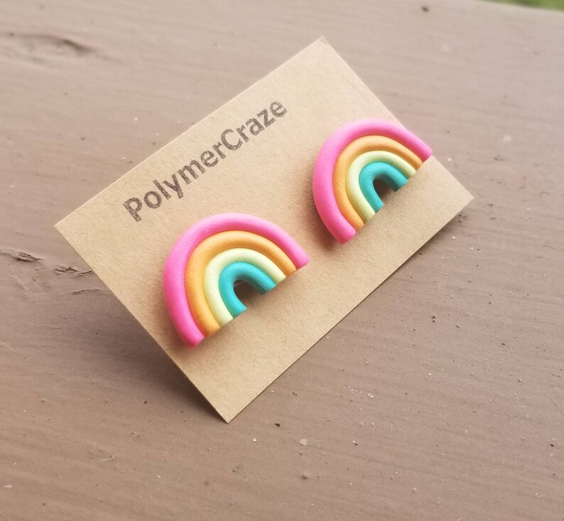 May include: A pair of rainbow-shaped stud earrings. Each earring features a pink outer band, followed by bands of orange, yellow, and turquoise. The earrings are displayed on a brown card with the text "PolymerCraze".