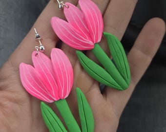 May include: A pair of handmade earrings featuring bright pink tulip flower designs with green stems and leaves. The earrings have silver-colored posts. The earrings are held in a hand.