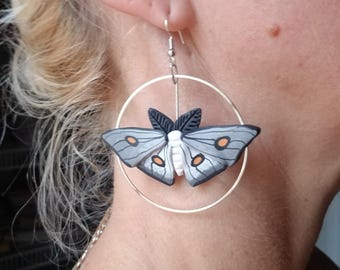 May include: Silver hoop earrings with a gray and black moth charm. The moth has white accents and orange markings on its wings.