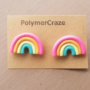 May include: A pair of rainbow-shaped stud earrings. Each earring features a pink outer layer, followed by orange, yellow, and turquoise layers. The earrings are displayed on a brown card with the text "PolymerCraze".