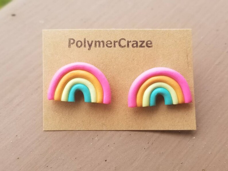 May include: A pair of rainbow-shaped stud earrings. Each earring features a pink outer layer, followed by orange, yellow, and turquoise layers. The earrings are displayed on a brown card with the text "PolymerCraze".
