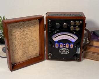 Pode incluir: Um medidor elétrico Weston vintage alojado numa caixa de madeira polida com tampa articulada. A face preta apresenta um indicador analógico curvo e quatro tubos nixie de brilho azul que mostram 12:00. A tampa interior contém uma folha de instruções.