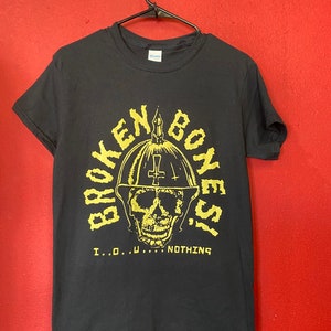 May include: Black t-shirt with a yellow graphic of a skull wearing a helmet with a cross on top. The text "BROKEN BONES" is written around the skull and "I...O...U...NOTHING" is written below the skull.