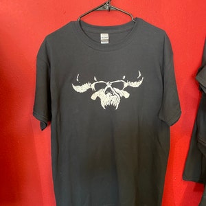 May include: Black t-shirt with a white skull graphic. The skull has horns and fangs. The t-shirt is hanging on a hanger against a red wall.