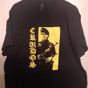 Puede incluir: Camiseta negra con un gráfico amarillo de una persona sosteniendo un rifle, con la palabra "CRUOS" en letras góticas amarillas. El diseño es una ilustración audaz y estilizada.