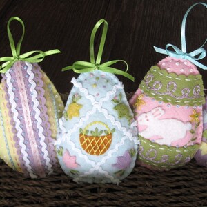 May include: Five felt Easter egg ornaments with different designs. The ornaments are decorated with embroidered details, including a chick, a basket, a bunny, and hearts. The ornaments are hung with ribbon loops.