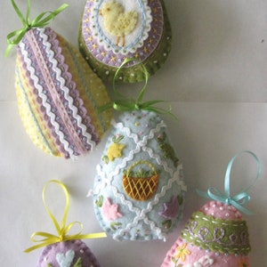 May include: Five felt Easter egg ornaments with different designs. The ornaments are decorated with embroidered details, including a chick, a basket, a bunny, and hearts. The eggs are in shades of green, pink, blue, and yellow.