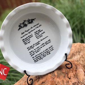 May include: White ceramic pie plate with a black handwritten pumpkin pie recipe. The recipe includes ingredients like pumpkin, eggs, milk, and spices. The plate is on a black metal stand.