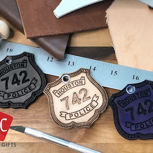 Könnte beinhalten: Drei Leder-Polizeibadges mit dem Text "HOUSTON 742 POLICE" in Schwarz, Braun und Lila. Die Abzeichen befinden sich auf einer Holzoberfläche mit einem Lineal und Lederresten.