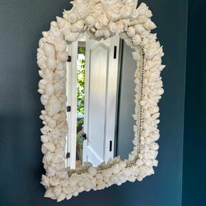 May include: An arched mirror with a frame entirely covered in various white seashells. The mirror is hanging on a dark blue wall. The reflection shows a white door and greenery.