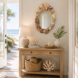 May include: An oval wall mirror framed with a variety of natural seashells in shades of white, tan, and pink. The mirror is mounted on a white paneled wall above a light wood console table featuring two drawers and a lower shelf for storage.