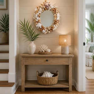 May include: An oval mirror framed with various seashells hangs above a wooden console table. The table holds a white vase with a palm frond, a white coral piece in a woven tray, and a lamp with a woven shade. A woven basket with a large shell sits below.