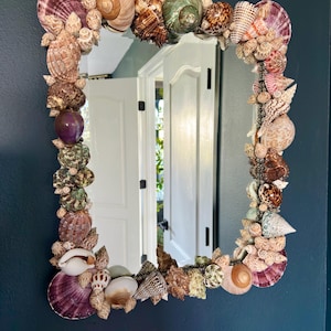 May include: A rectangular mirror with a frame intricately decorated with various seashells in shades of brown, tan, purple, and green. The mirror is mounted on a dark blue wall, reflecting a white door and natural light.