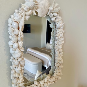 May include: An arched mirror with a frame densely decorated with various white seashells. The mirror reflects a bedroom interior. The seashells are of different sizes and shapes, creating a textured, coastal-themed decorative piece.