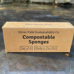 May include: A brown cardboard box with the text "100% Plastic Free" and "Silver Falls Sustainability Co." printed on the top. The box also has the text "Compostable Sponges" printed in large letters. Below the text, there are two smaller boxes with the text "Set of 4" and "Made in the USA".