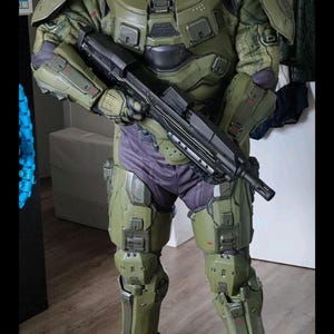 Könnte beinhalten: Ein olivgrüner und grauer Panzeranzug mit goldenem Visier, der an eine Science-Fiction-Figur erinnert. Das Kostüm umfasst einen Helm, Schulterpolster, Brust- und Beinpanzer sowie ein schwarzes Gewehr. Die Person steht auf einem Holzboden.