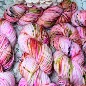 May include: Close-up of several skeins of hand-dyed yarn in shades of pink, white, orange, and green. The yarn is arranged on a floral patterned fabric, with a cluster of white paper flowers in the background. The yarn appears soft and textured.
