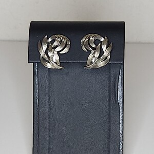 Puede incluir: Pendientes plateados con forma de hojas, presentados en un estuche de joyería de polipiel negra. Los pendientes tienen un acabado texturizado y un diseño en capas. El estuche rectangular está cerrado, con una superficie brillante.