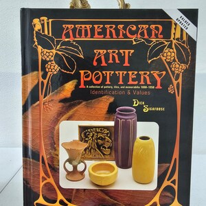 May include: A book titled "American Art Pottery: A collection of pottery, tiles, and memorabilia 1880-1950 Identification & Values" by Dick Sigafoos. The cover features a decorative design with a lion and several pieces of pottery in various colors.