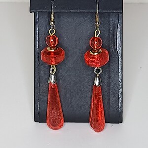 Puede incluir: Un par de pendientes rojos colgantes con una parte inferior en forma de lágrima y una parte superior redonda y facetada.