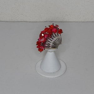 Op de afbeelding: Een zilveren ring met een cluster van rode en roze kralen. De ring staat op een witte displaystandaard.