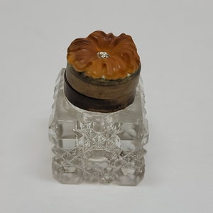 May include: A clear glass bottle with a square base and a cut glass design. The bottle has a brown wooden lid with a carved orange flower on top.