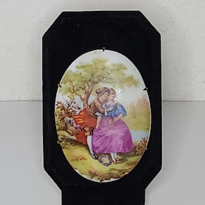 Könnte beinhalten: Eine ovale Porzellanplatte mit einer bemalten Szene eines Paares in einer Gartenlandschaft. Die Platte ist in einem schwarzen achteckigen Rahmen gerahmt.