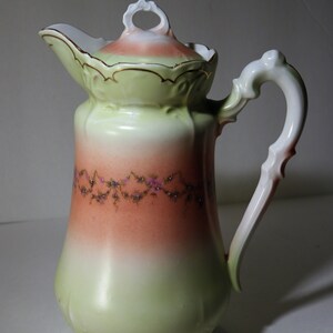 May include: A vintage porcelain creamer with a green and pink ombre glaze. The creamer has a delicate floral design and a gold trim around the lid.