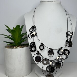 May include: A layered necklace with multiple black metal circles and beads. The necklace is displayed on a white mannequin bust.