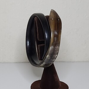 May include: Two bracelets on a brown velvet display stand. One bracelet is a solid black, the other is a curved horn shape with a light brown and white pattern. The stand has a round base and a cone-shaped top.
