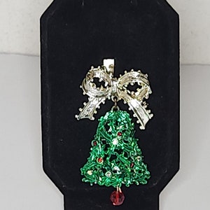 May include: A green Christmas tree-shaped bell with red and white rhinestones, topped with a gold bow with white rhinestones. The bell is hanging from a red rhinestone.